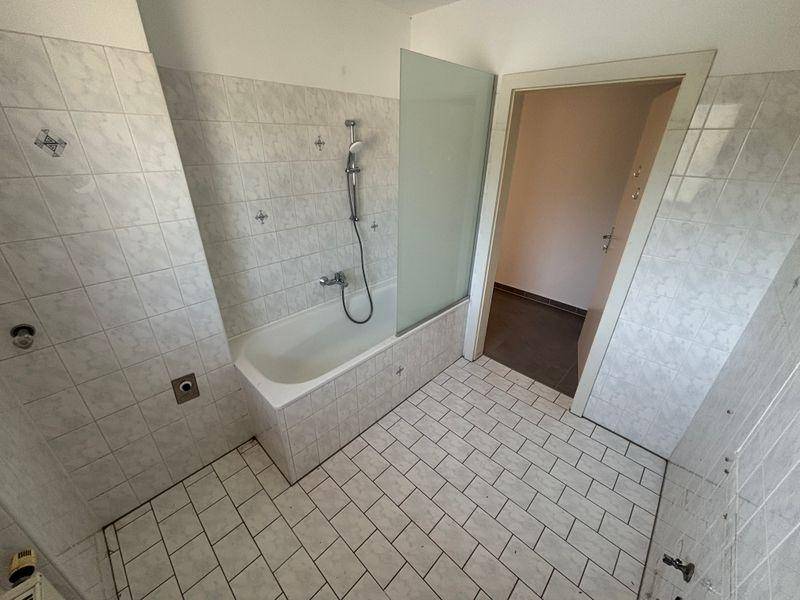 Helles Badezimmer mit Badewanne, Duschvorrichtung und weißen Fliesen an Wänden und Boden.