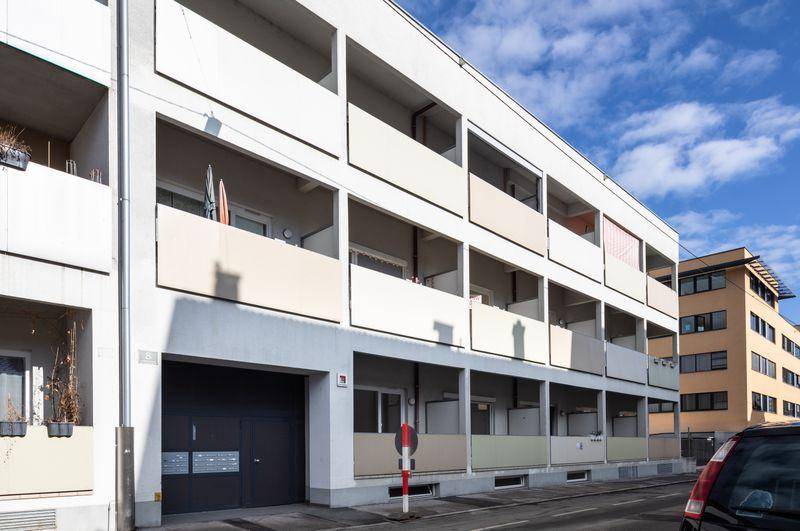 Mehrfamilienhaus mit moderner weißer Fassade, mehreren Balkonen und integrierter Tiefgarage an einer Straße.