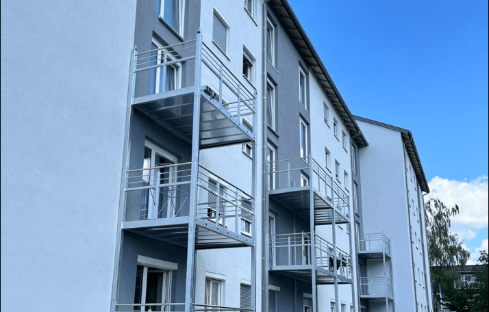 Moderne Fassade eines Mehrfamilienhauses mit mehreren Balkonen, großen Fenstern und Rollläden unter blauem Himmel.