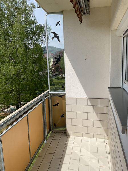 Balkonecke mit Fliesenboden und dekorativen Vogelsilhouetten auf einer Glasscheibe, umgeben von Grün.
