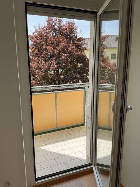 Blick von einem Innenraum durch eine offene Balkontür auf einen gefliesten Balkon mit Baum.