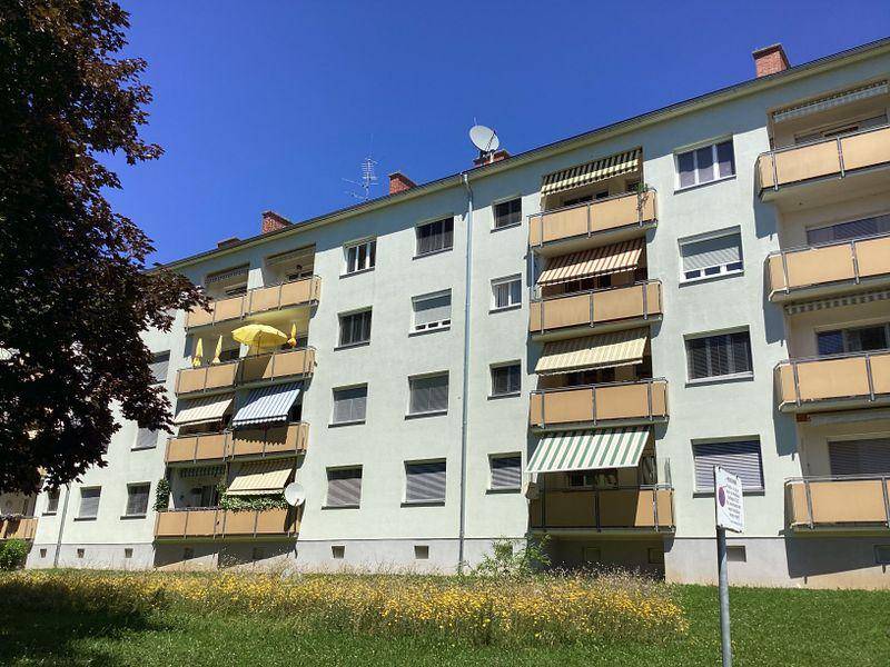 Mehrfamilienhaus mit zahlreichen Balkonen, einigen Markisen und einer grünen Rasenfläche im Vordergrund.