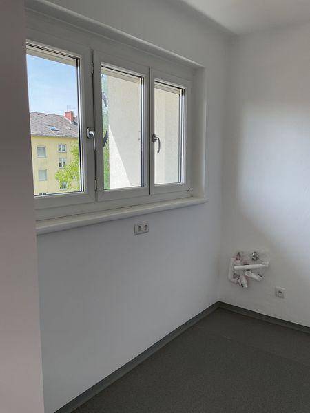 Leerer Raum mit grauem Boden, Fenster und Anschlüssen für eine zukünftige Küche.