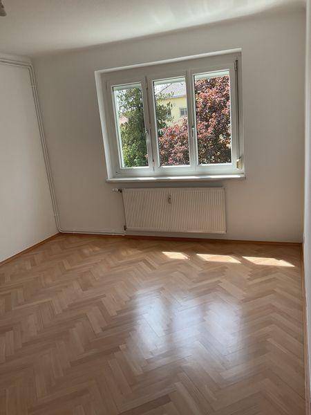 Heller Raum mit Fischgrätparkett, Fenster und Heizkörper, der viel Tageslicht hereinlässt.