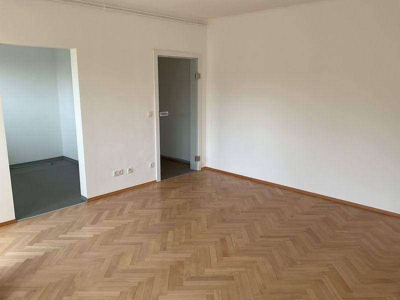 Geräumiger Raum mit Fischgrätparkett und Zugang zu einem angrenzenden Zimmer mit grauem Boden.