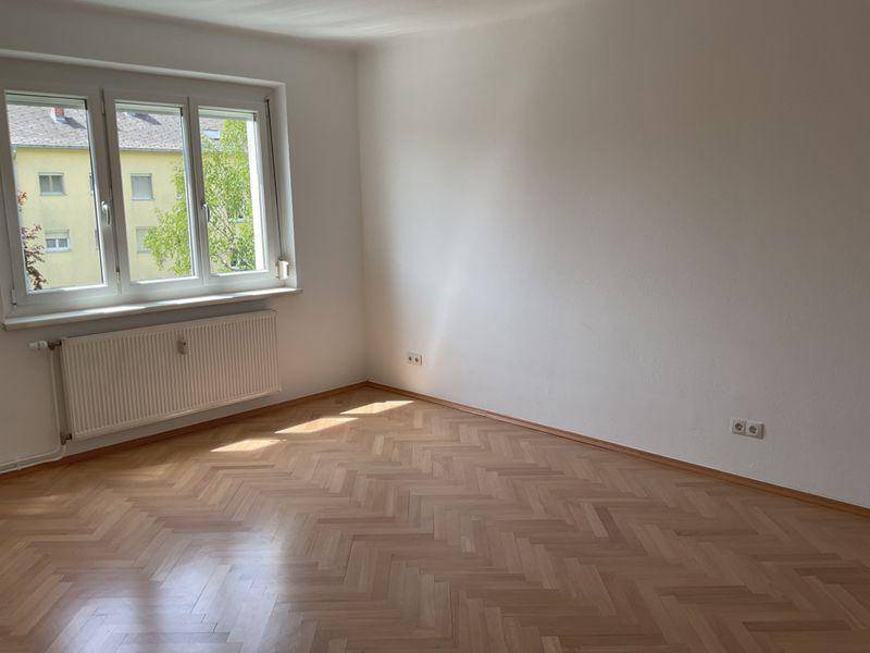 Geräumiger Raum mit Fischgrätparkett, Fenster und Heizkörper, mit Blick auf ein Nachbargebäude.