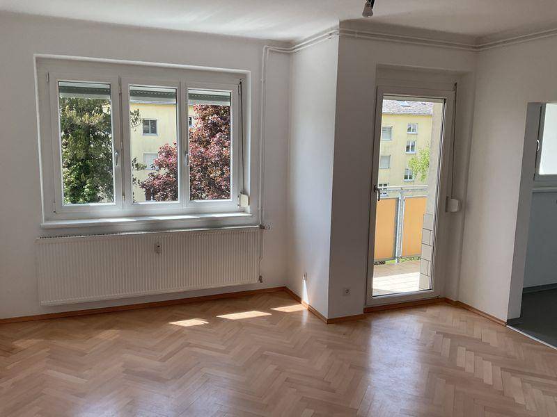 Lichtdurchfluteter Wohnraum mit Fischgrätparkett, Fenster und Balkonzugang, mit Blick ins Grüne.