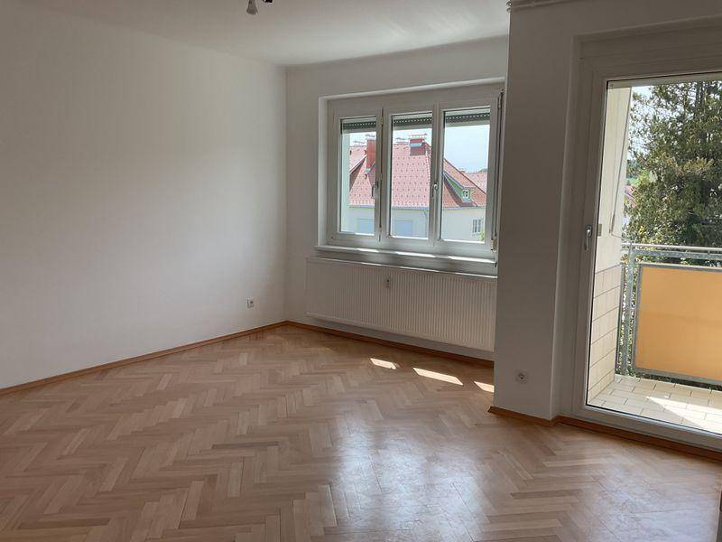 Heller Wohnraum mit Fischgrätparkett, Fenster, Heizkörper und direktem Zugang zum Balkon.