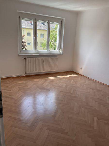 Lichtdurchfluteter Raum mit Fischgrätparkett, Fenster und Heizkörper, mit Blick auf die Umgebung.