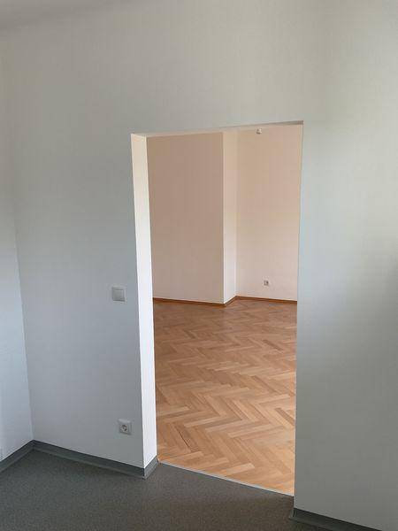 Blick vom Flur mit grauem Boden in einen Raum mit hellem Fischgrätparkett.