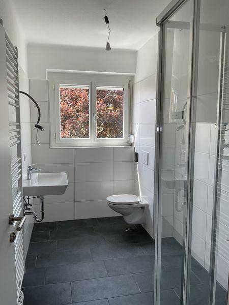 Modernes Badezimmer mit dunklen Fliesen, Duschkabine, wandhängendem WC und Fenster.