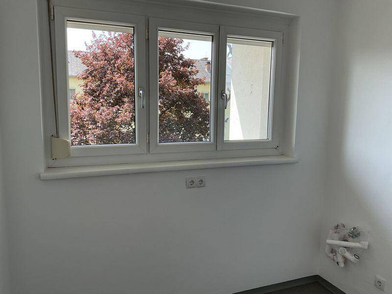 Heller Raum mit grauem Boden und Fenster, das einen Blick auf einen Baum mit roten Blättern bietet.