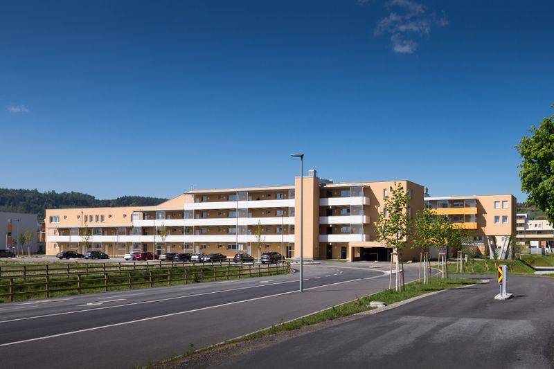 Modernes Mehrfamilienhaus mit hellbeiger Fassade, zahlreichen Balkonen und Außenparkplätzen unter blauem Himmel.