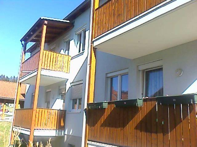Mehrfamilienhaus mit mehreren Balkonen und Holzverkleidung, umgeben von grüner Landschaft.