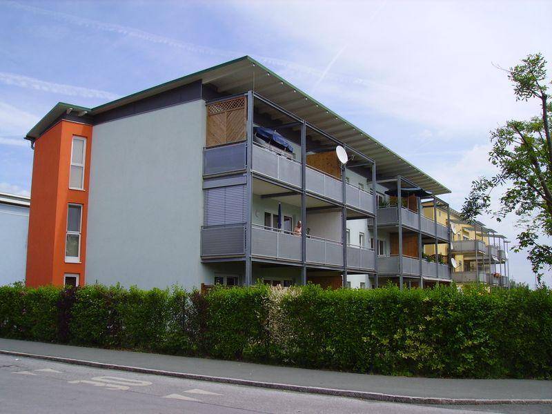 Ansicht eines modernen Wohngebäudes mit mehreren Balkonen und einer grünen Hecke entlang der Straße.