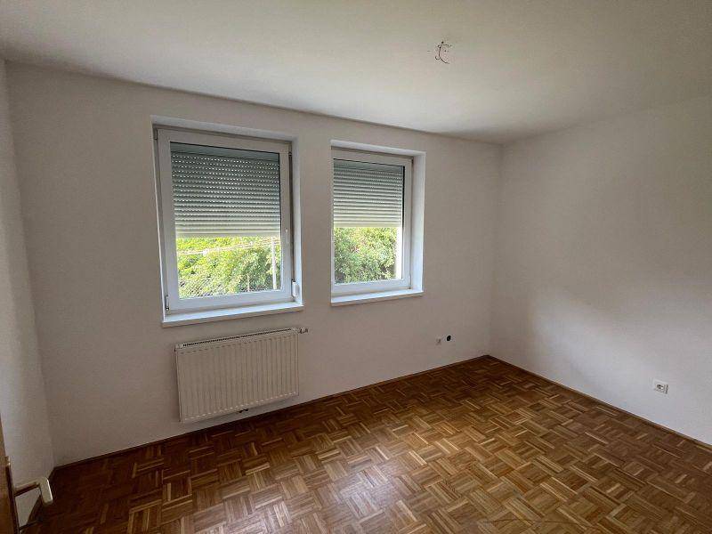 Helles Zimmer mit Parkettboden, weißen Wänden und zwei Fenstern mit Blick ins Grüne.
