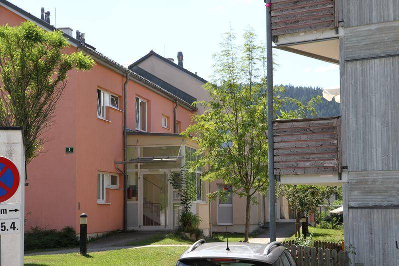 Mehrfamilienhaus mit lachsfarbener Fassade, umgeben von Grünflächen und Bäumen, mit Bergblick im Hintergrund.