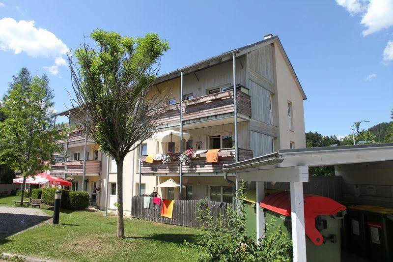 Mehrfamilienhaus mit Balkonen, umgeben von Grünflächen und Bäumen unter blauem Himmel.
