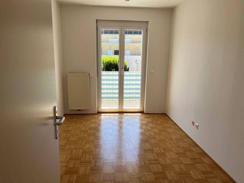 Schlafzimmer mit hellem Parkettboden, weißen Wänden und Fenster mit Sichtschutz zum Außenbereich.