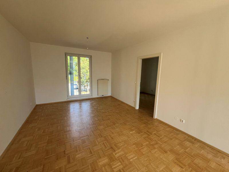 Helles, leeres Wohnzimmer mit Fischgrätparkett, weißen Wänden und Zugang zu einem Balkon.