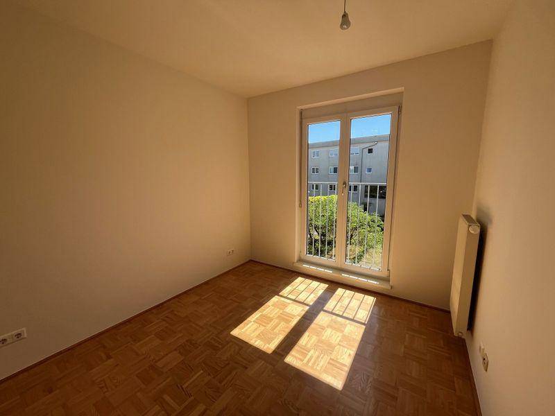 Helles Schlafzimmer mit Parkettboden, weißen Wänden und sonnigem Ausblick auf die Umgebung.