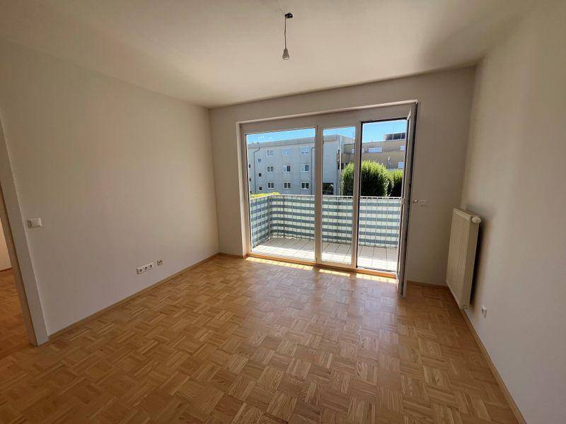 Geräumiges Wohnzimmer mit Parkettboden, weißen Wänden und breitem Zugang zum Balkon.