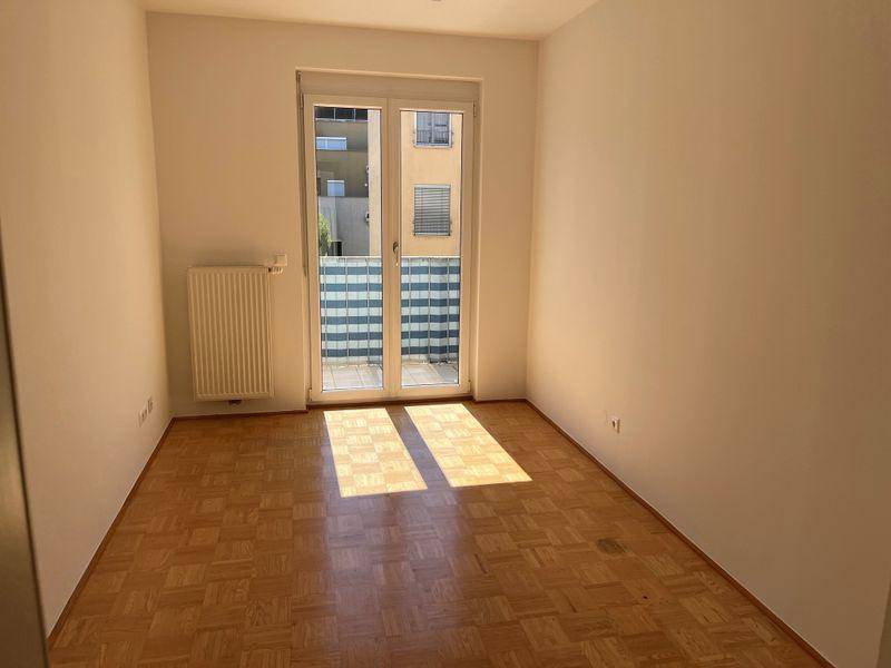 Sonniger Raum mit Parkettboden, weißen Wänden und viel Tageslicht durch die große Balkontür.