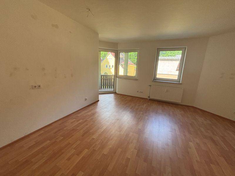 Helles, leeres Zimmer mit Laminatboden, Heizkörper und Zugang zu einem Balkon.