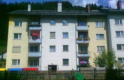 Mehrfamilienhaus mit Balkonen und einem kleinen Spielbereich im Vordergrund, umgeben von Bäumen.