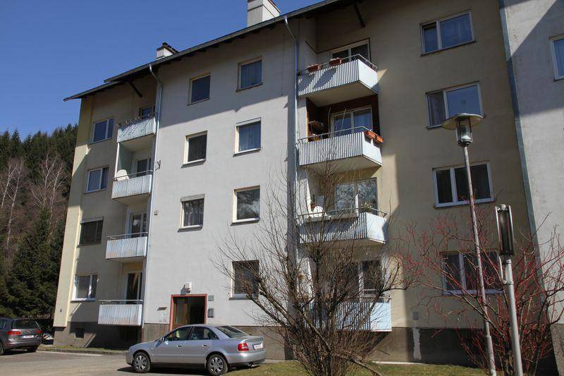 Wohngebäude mit heller Fassade, Balkonen und geparkten Autos, umgeben von Bäumen und einem Hügel.