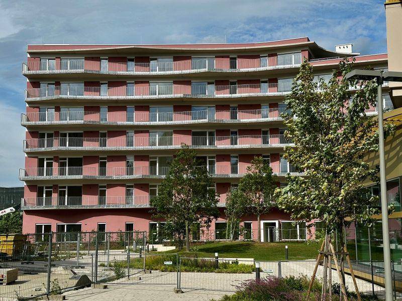 Ansicht der modernen Wohnanlage mit zahlreichen Balkonen und junger Bepflanzung vor der roten Fassade.