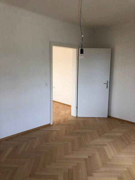 Zimmer mit Fischgrätparkett und Zugang zu einem weiteren Raum, hell und offen gestaltet.