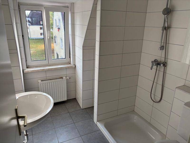 Badezimmer mit Dusche, Waschbecken, WC, weißen Fliesen und einem Fenster mit Heizkörper.