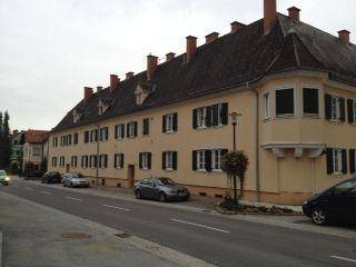 Seitenansicht eines historischen Gebäudes mit Erker und Mansarddach entlang einer Straße mit parkenden Autos.