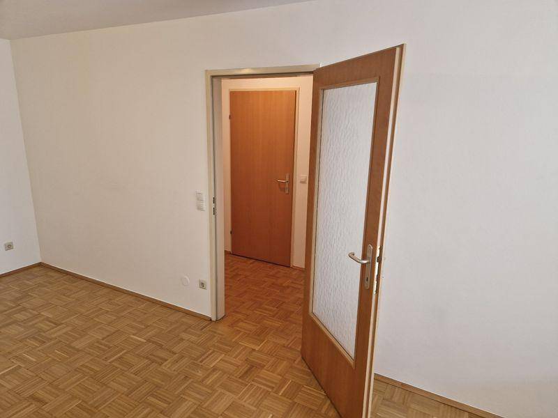 Geräumiger, leerer Wohnraum mit Parkettboden und einer Tür mit Glaseinsatz.