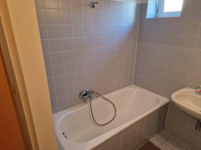 Badezimmer mit grauen Fliesen, einer Badewanne und einem kleinen Waschbecken unter einem Fenster.
