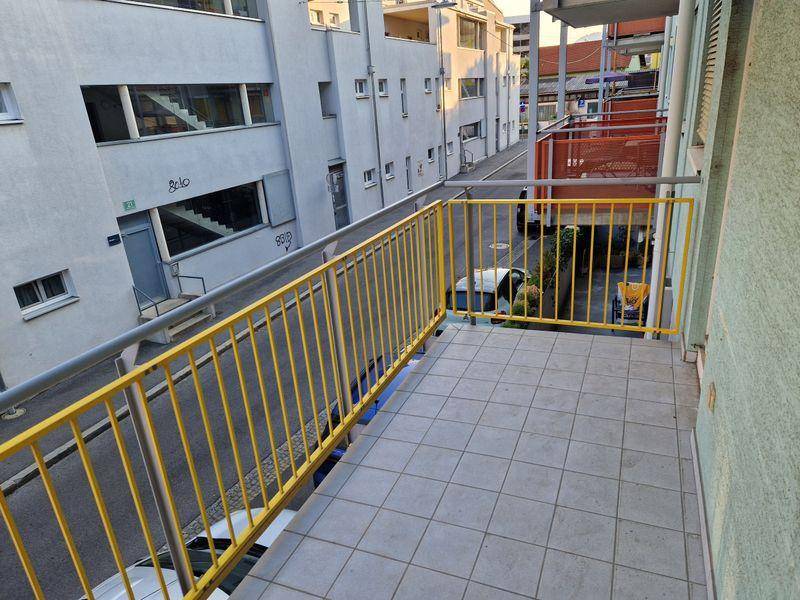 Balkon mit gelbem Metallgeländer und Fliesenboden, umgeben von städtischer Architektur.