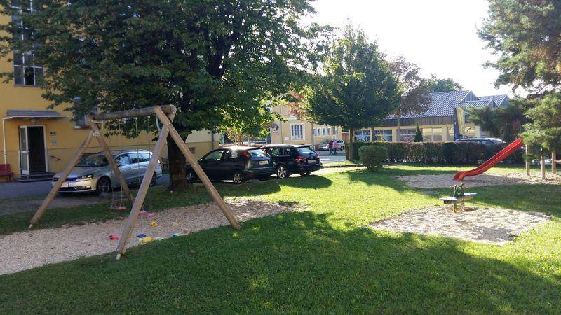 Grüner Spielplatz mit Schaukel und Rutsche, umgeben von Bäumen und Parkplätzen.