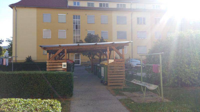 Rückseite des Gebäudes mit überdachtem Müllbereich und gepflegter Hecke im Vordergrund.