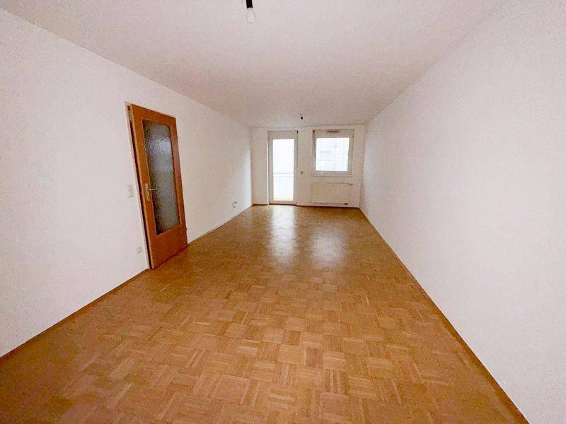 Geräumiges, leeres Wohnzimmer mit Parkettboden und Zugang zu einem Balkon.