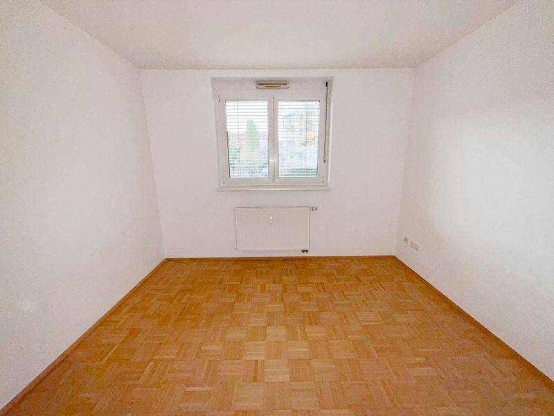 Leeres Schlafzimmer mit Parkettboden und einem Fenster, das viel Tageslicht hereinlässt.