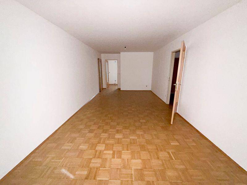Langes, leeres Wohnzimmer mit Parkettboden und Zugang zu weiteren Räumen.