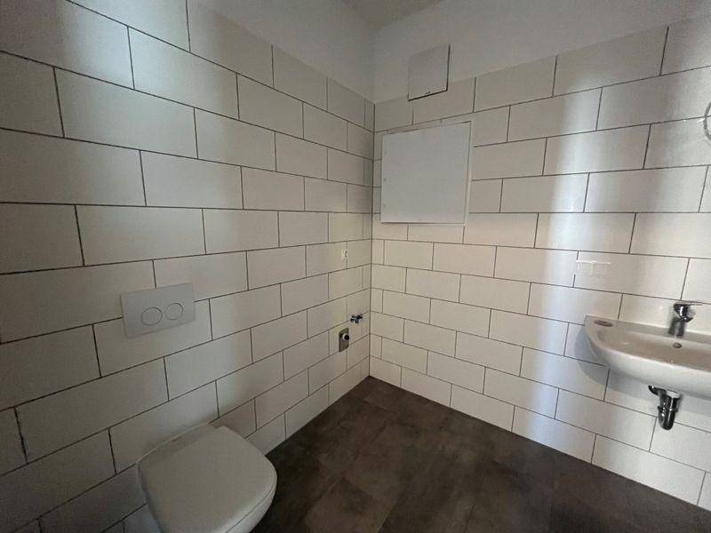Separates WC mit weißen Wandfliesen, wandhängender Toilette und kleinem Waschbecken.