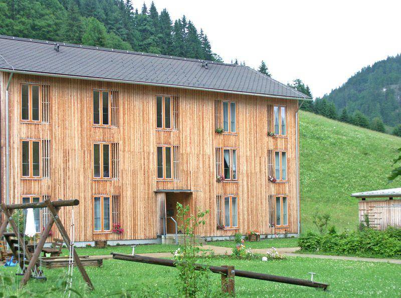 Modernes Holzhaus mit Satteldach und großzügigen Fenstern, umgeben von grüner Natur und einem Spielplatz.