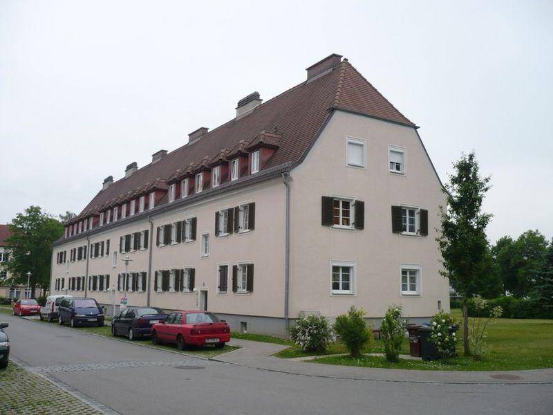 Langes Wohngebäude mit heller Fassade, Ziegeldach und Fensterläden, gelegen an einer Straße mit parkenden Autos und Grünflächen.