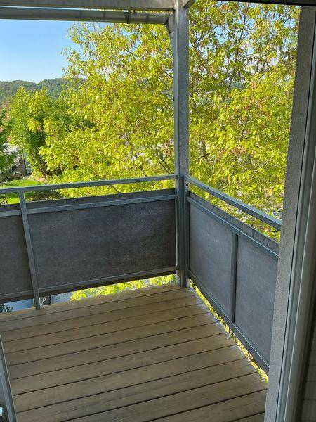 Heller Balkon mit Holzboden und modernem Metallgeländer, umgeben von grünen Bäumen.