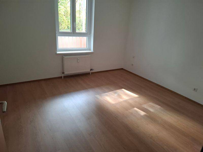 Leerer Raum mit Laminatboden, großem Fenster und weißem Heizkörper, ideal als Schlafzimmer.