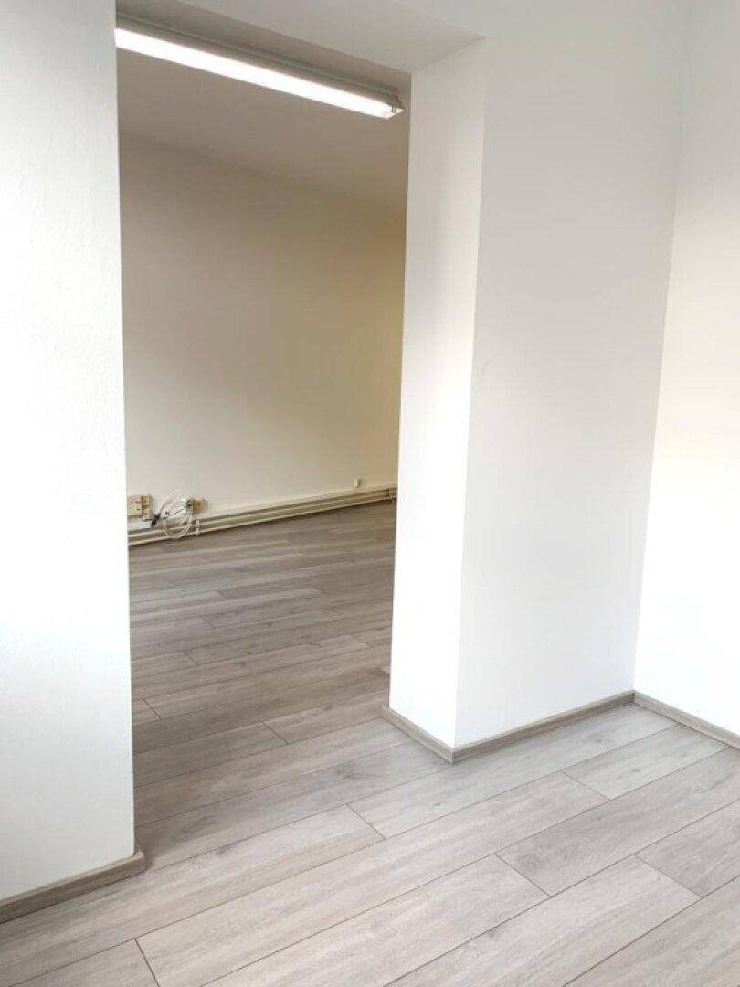 Blick durch einen Türrahmen in einen weiteren hellen Büroraum mit gleichem Laminatboden.