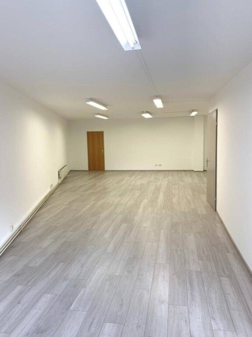 Geräumiger, leerer Büroraum mit hellem Laminatboden, weißen Wänden und mehreren Deckenleuchten.