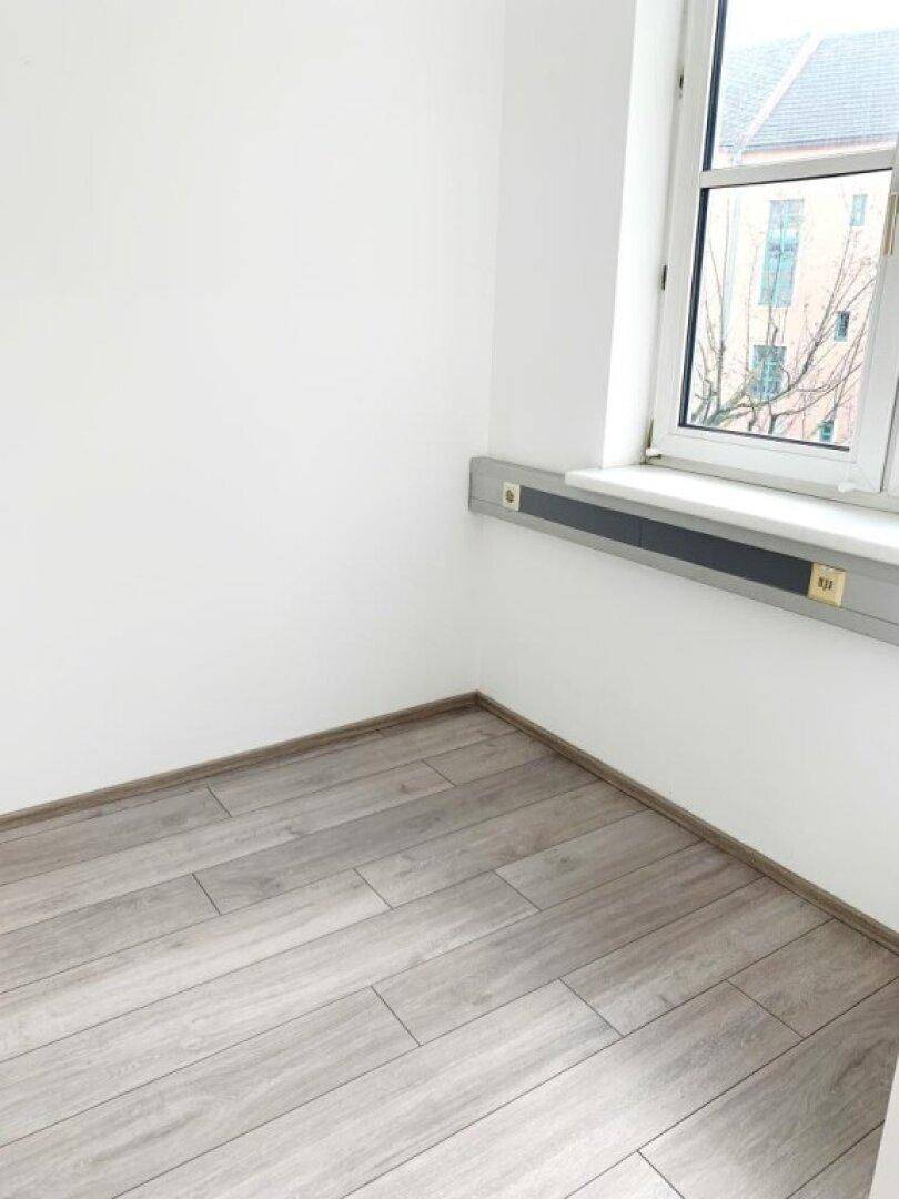 Helle Ecke eines Büroraums mit Fenster, hellem Laminatboden und weiß gestrichenen Wänden.